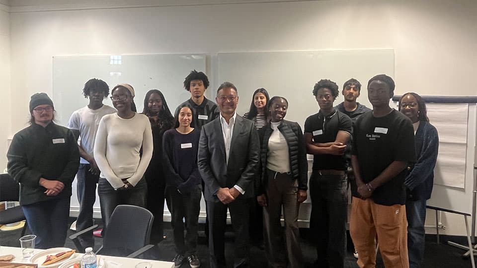 Gagan is wearing a grey suit and is smiling. He is standing with 12 people who attended the Lunch with a Leader talk.